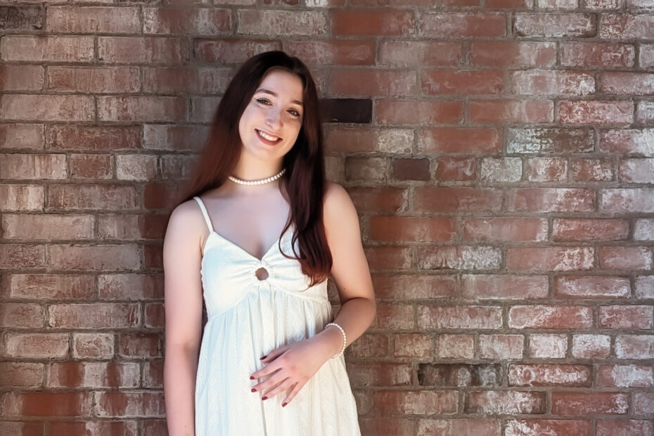 A teenaged girl posing in front of a brick wall wearing a white dress.
